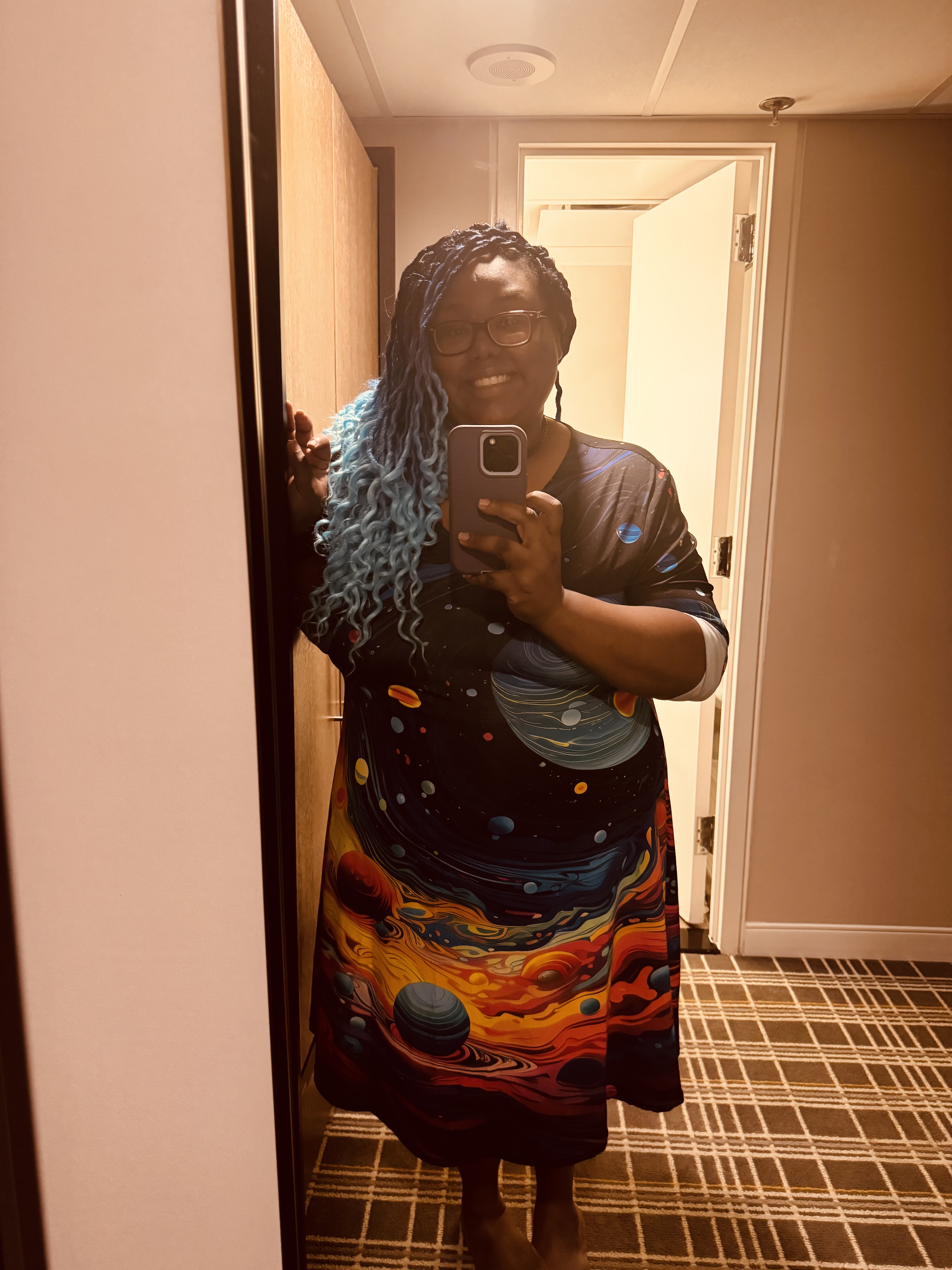 Me with ombre blue-tinted braids taking a mirror selfie while wearing a colorful galaxy themed dress with designs in red, orange, yellow, and other vibrant colors.