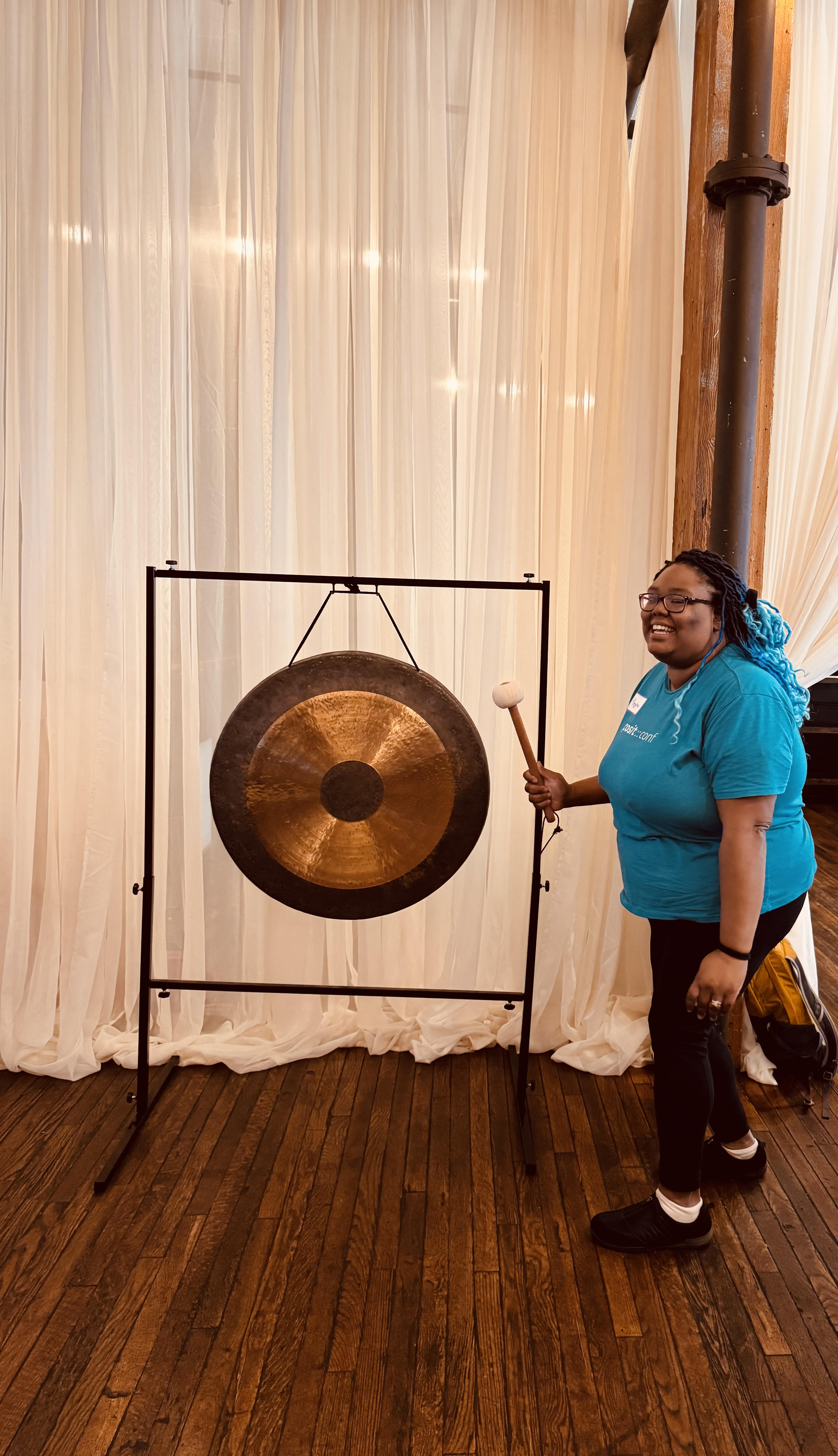 Me smiling with ombre blue-tinted hair wearing a turquoise posit conf shirt and glasses, holding a mallet next to a large bronze gong mounted on a black metal stand. The setting is indoors with white curtains and wooden flooring in the background.